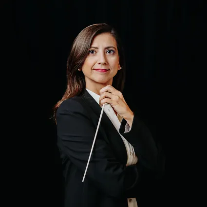 A woman in a black suit confidently holds a conductor's baton against a dark background. Her expression is focused and poised, conveying leadership.