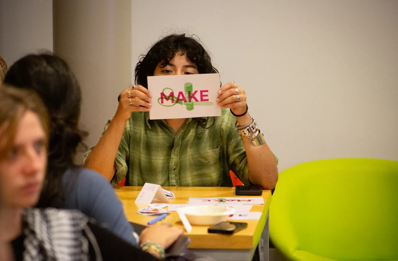 A student holds a paper with the words Make in front of their face while looking at the camera at ZineFest 2024