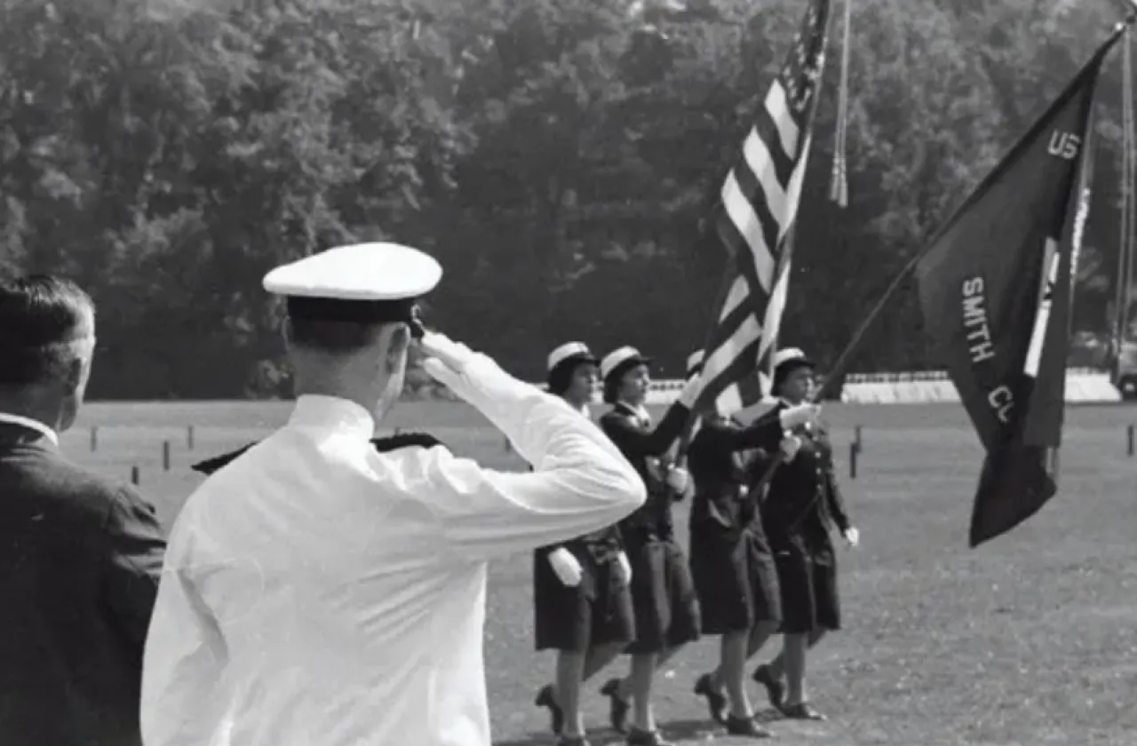 A male soldier salutes female soldiers as they march across the lawn carrying an American flag and a Smith College flag