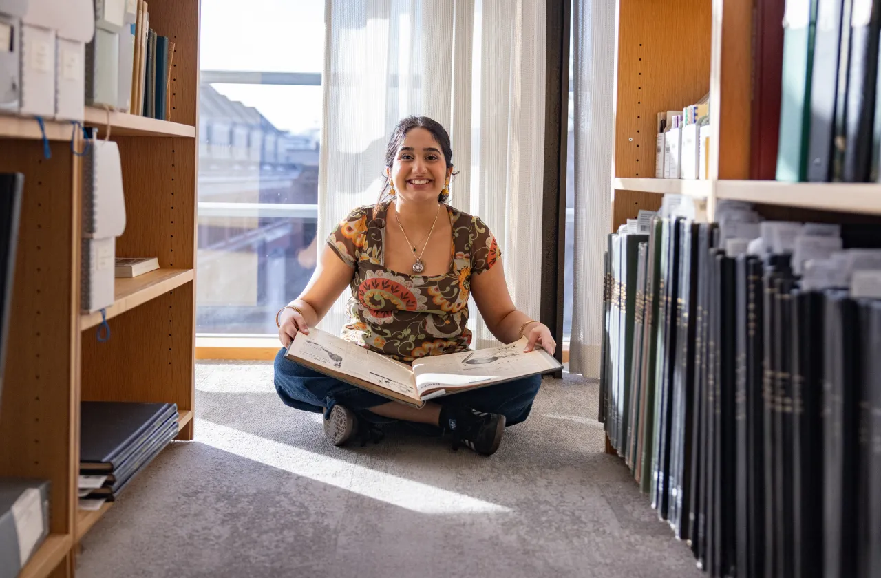 A student sitting on the floor of the library with an oversized book in her lap