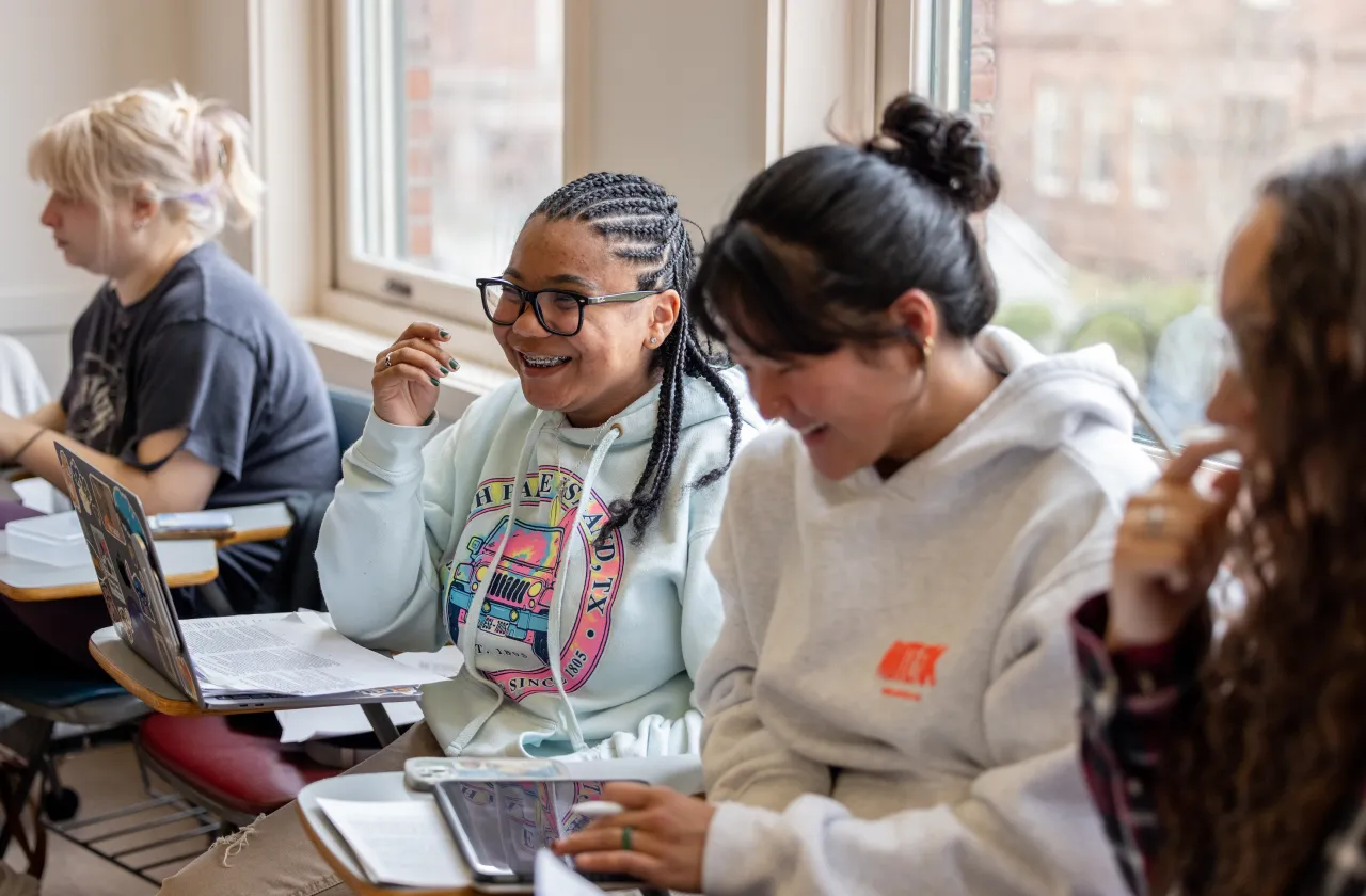 Students smile while seated at desks in front of laptops