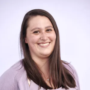 A woman with long brown hair smiles at the camera. She is wearing a light purple cardigan and blouse.