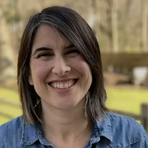Headshot of a smiling woman with shoulder-length dark hair showing subtle streaks of gray, wearing a blue denim button-down shirt. She is outdoors with a soft, blurred background featuring trees and a lawn.