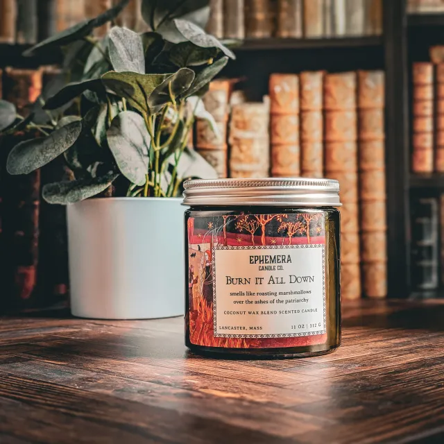 A candle sitting on a desk. The label says "Burn it all down, smells like roasting marshmallows over the ashes of the patriarchy."