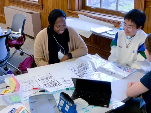 Three students working in a small group. The main figure is a young black woman in a headscarf.