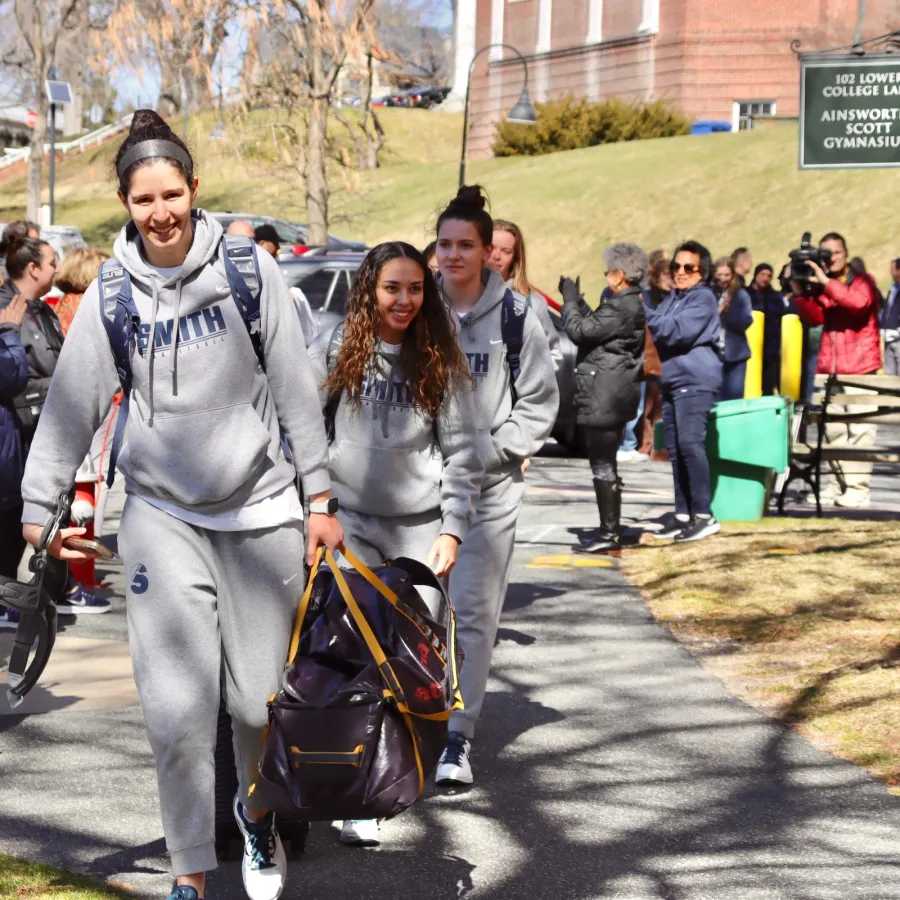 Basketball players, carrying luggage and smiling, walk along the path toward the bus