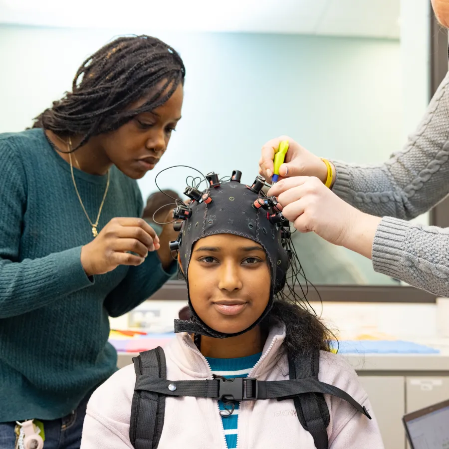Student researcher Kris Cheaye and lab manager Maddie Scricco fit an fNIRS cap to Gloriamar Esteves’s head