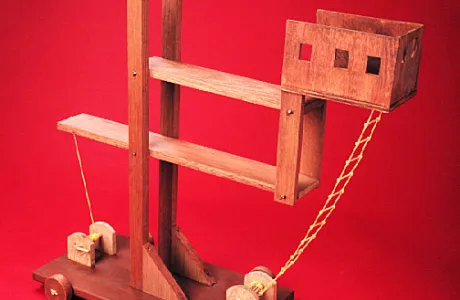 Wooden catapult model features beams, a basket on the end, rope details, and wheels