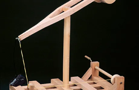 A wooden mechanical model with a geared lifting arm is displayed on a grid-like base, demonstrating basic engineering principles