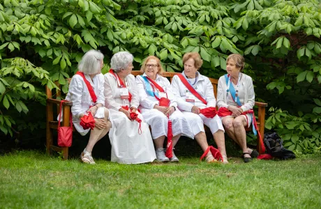Smithies in white wearing red sashes sit and chat on a bench