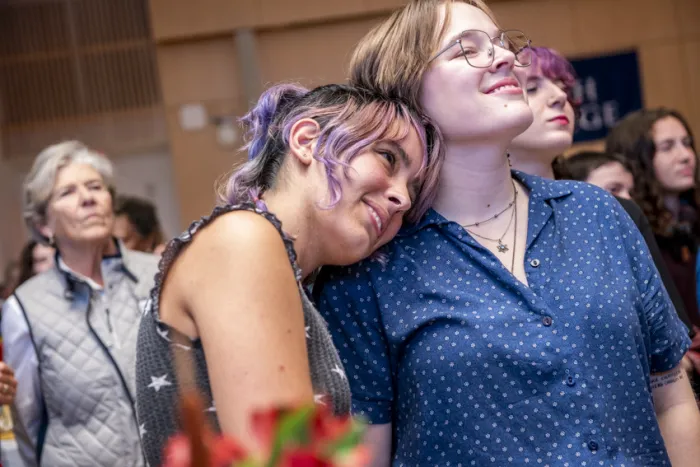 Two students, one with their head on the other's shoulder, smile and look on together at the festivities.