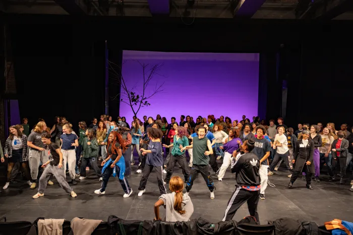 A large group of students on a stage in front of a purple backdrop, dancing as a group