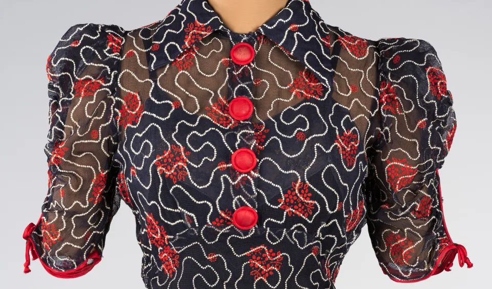 Photo of a sheer, button-up, navy blue, collared top with red buttons and elaborate pattern consisting of wavy white lines and red flowers.