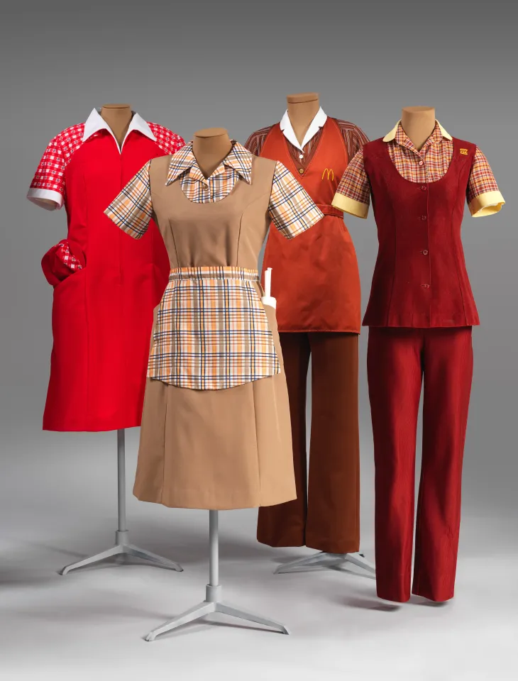 Four fast-food uniforms, ranging in color from red to beige, featuring aprons and button-up tops.