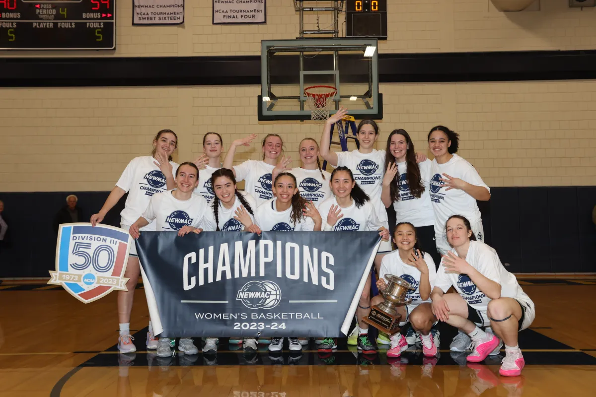 The 2023-2024 basketball team posing with their NEWMAC championship banner.