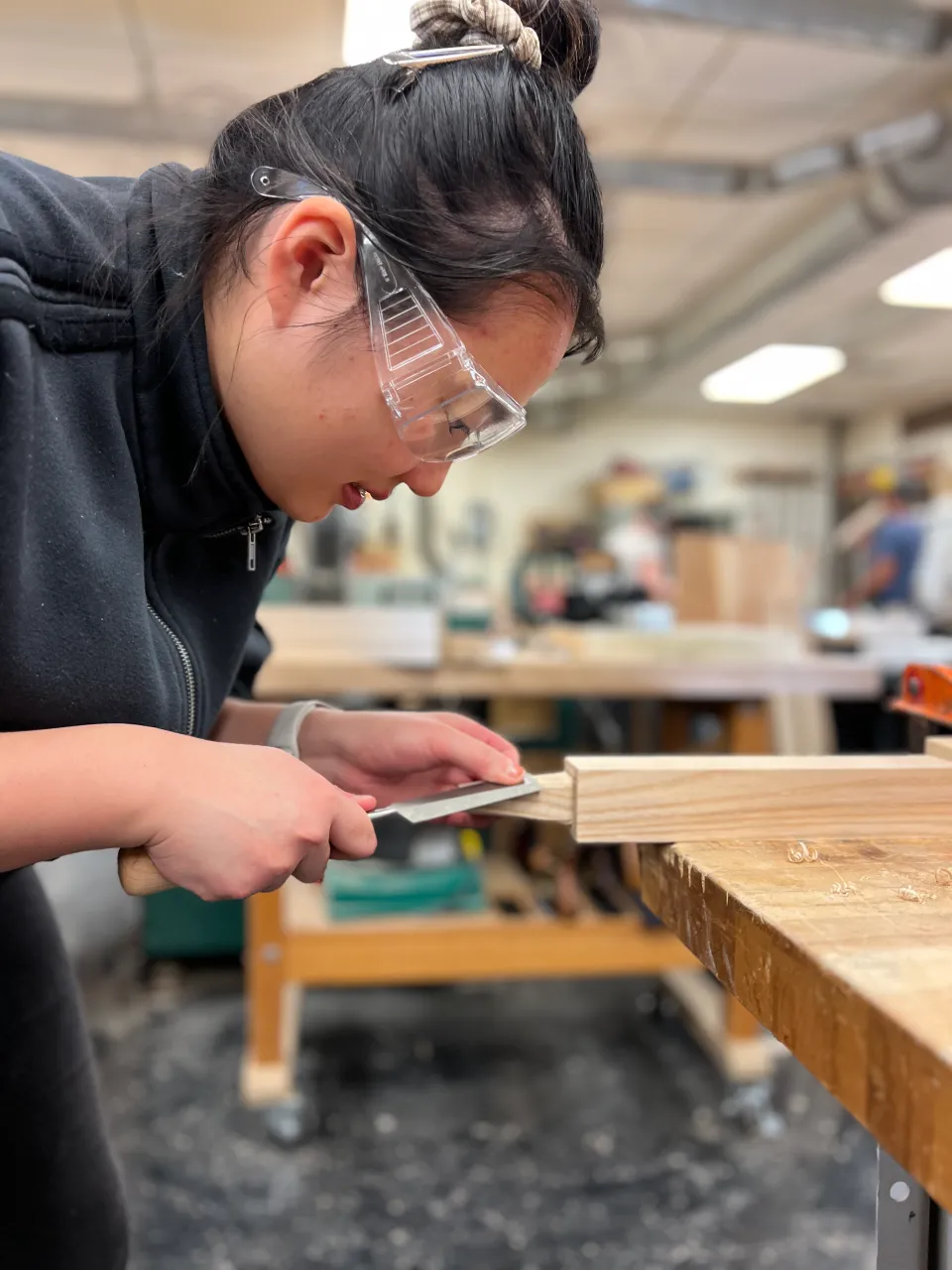 A student in a woodworking class, wearing safety glasses, files the tenon on a piece of ash wood